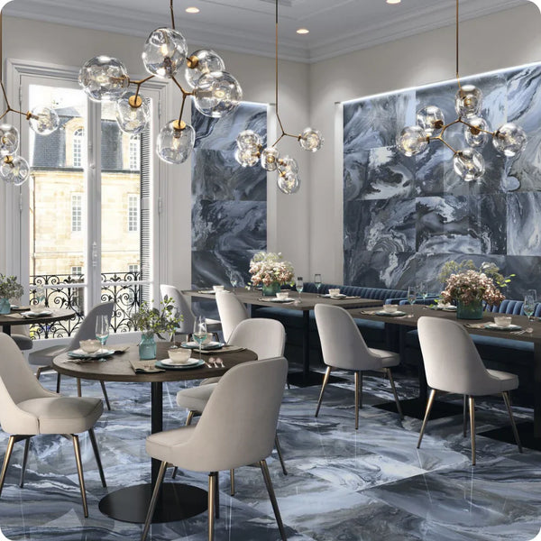 Elegant restaurant interior featuring marble-look flooring and wall panels, modern round tables, upholstered chairs, floral centerpieces, and artistic bubble chandeliers with large windows letting in natural light.